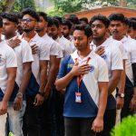 Line of young men in matching white and blue polo shirts with orange lanyards, standing with right hands over their hearts outdoors, likely during a pledge or oath.