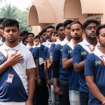 Young men in blue-and-white polo shirts with orange lanyards stand in a line outside, many with hands over their hearts in a pledge gesture.